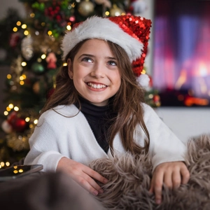 Little girl wearing red sequined Christmas hat with fuzzy pom-pom in warm holiday setting