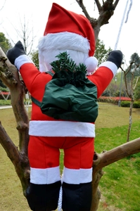 Outdoor detail of a plush Santa Claus climbing a white rope with a green gift bag on a tree