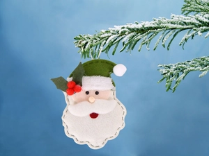 Dettaglio di un addobbo in feltro di Babbo Natale con barba bianca su un ramo di pino innevato
