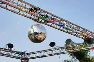 Disco mirror ball hanging from a stage truss with colorful flags and clear blue sky