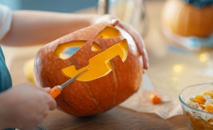 Action shot of hands using an orange pumpkin carving saw to cut festive patterns on a pumpkin