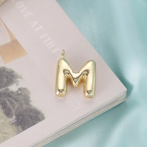 Polished gold bubble letter M pendant on a soft pink and turquoise silk background