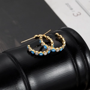 Close-up of golden C-shaped evil eye earrings with blue enamel and clear zircons