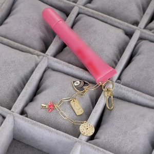 Detalle de brillo de labios rosa con dijes de ojo y hongo dorados sobre joyero gris