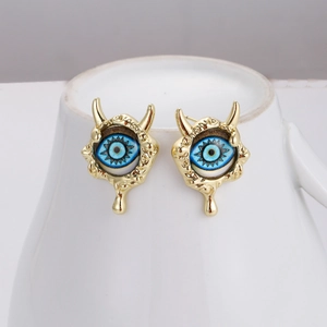 Close-up of golden devil eye stud earrings with blue pupils on white ceramic