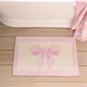 Cute pink bow absorbent bath mat with plush texture on light wood floor next to a bathtub
