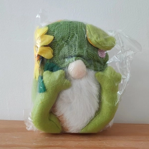 Detail eines grünen gestrickten Froschzwergs mit Sonnenblumenhut in Verpackung