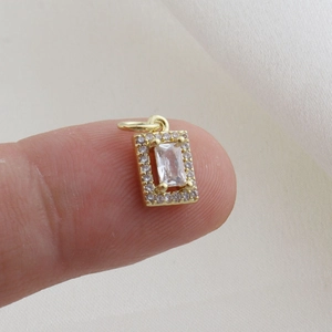 Close-up of a small gold rectangular pendant with clear zircon held on a fingertip