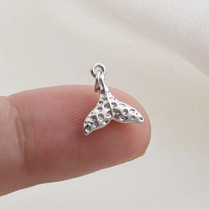 Detail of silver whale tail charm with textured surface held on fingertip for scale