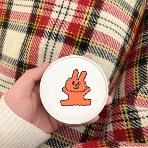Close-up of cute orange rabbit sticker on a round surface held by hand against plaid fabric
