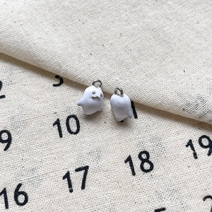 Détail de deux breloques fantômes en émail blanc avec boucles en argent sur lin