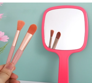 Detalle de un espejo de maquillaje rosa de mano con dos pinceles sobre fondo verde