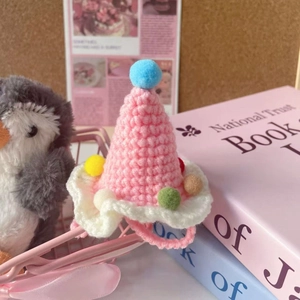 Pink and white knitted pet party hat with colorful pompoms shown in a cozy setting