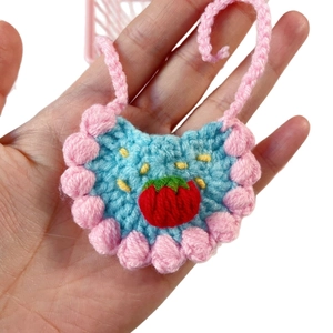 Handmade light blue knit doll bib with a red strawberry and pink pompom trim details