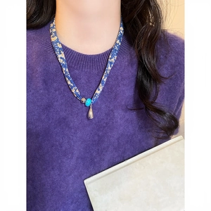 Primo piano di una collana in tessuto floreale blu con pendente a goccia su maglia viola.