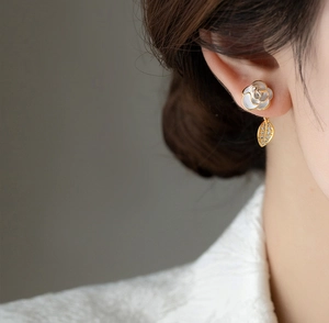 Close-up of elegant white camellia stud earring with gold leaf drop on model's ear.