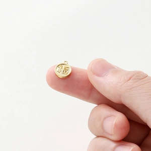 Macro detail of a tiny 18K gold-plated Fu pendant on a fingertip for size reference