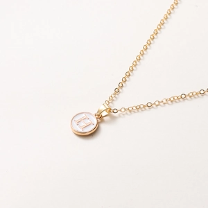 Détail macro du collier doré montrant la lettre H sur un pendentif en émail blanc