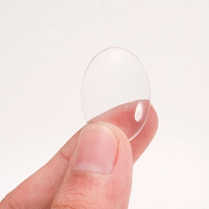 Transparent oval glass cabochon for DIY jewelry held by hand against a white background