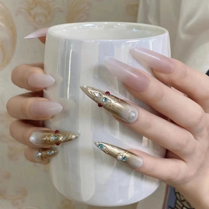 Macro view of long almond press-on nails with golden relief, pearls and colorful gemstones