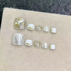 Faux ongles de pieds blancs faits main avec perles et détails dorés sur fond de velours