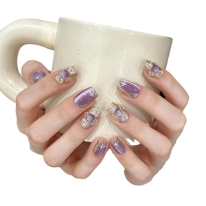Purple cat eye press-on nails with pearls and gold accents held against a ceramic cup