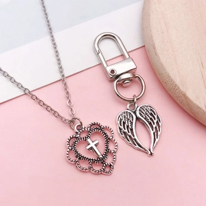 Collana cuore croce argento antico e ciondoli ali d'angelo su uno sfondo rosa morbido