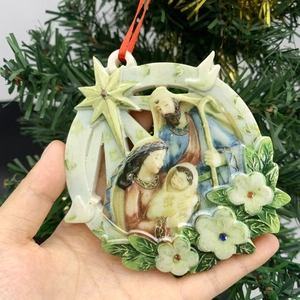 Close-up of Nativity scene Christmas ornament held in hand against green tree