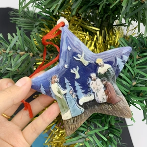 Detail of star-shaped resin Nativity ornament with red ribbon held by a Christmas tree