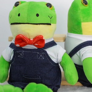 Green plush frog doll in denim overalls and red bowtie on a wooden bench