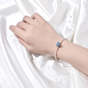 Detalle de pulsera de malla plateada con dije de corazón azul sobre fondo de seda blanca
