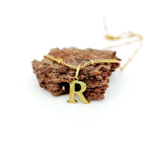 Détail d'un pendentif lettre R doré sur une chaîne à billes posée sur une roche volcanique