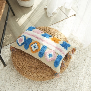 Housse de coussin bohème à pompons colorés posée sur un pouf en paille tressée