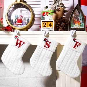 Three white cable knit Christmas stockings with red embroidered letters on a fireplace