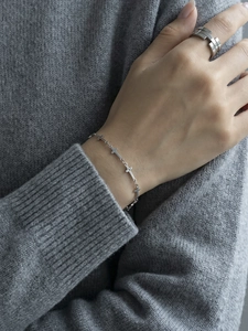Detail of silver cross charm bracelet and geometric layered ring on a grey knit sleeve