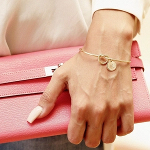Golden knot cuff bracelet with letter K charm held against a pink leather handbag