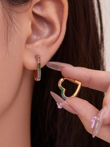 Detailed close-up of a heart-shaped gold earring with rainbow zircon stones on model's ear