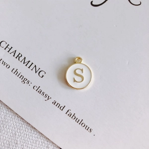 Dettaglio del ciondolo rotondo dorato con lettera S in smalto bianco su carta