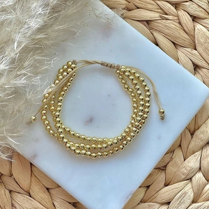 Drielagige vergulde kralenarmband met verstelbaar beige koord op marmeren dienblad
