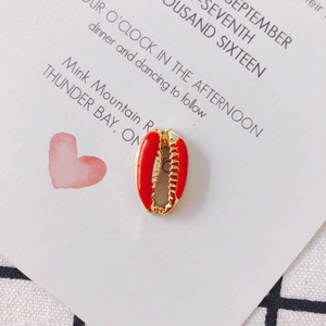 Macro detail of gold-plated cowrie shell pendant with red enamel inlay on white card