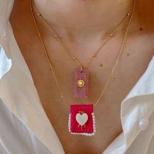 Layered gold necklaces featuring pink stone pendant and woven heart charm with pearls