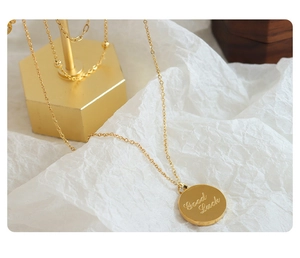 Close-up of gold layered coin necklace featuring an engraved Good Luck charm on a stand