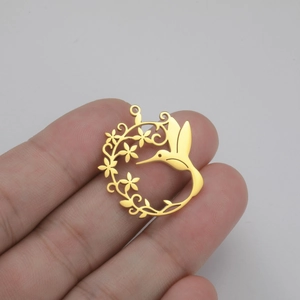 Pendentif fleur et colibri en acier inoxydable doré tenu entre les doigts sur fond gris
