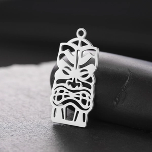 Detailed view of a silver hollow Tiki mask pendant on a dark pebble and stone tile