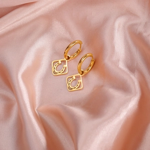 Close-up of golden stainless steel hoop earrings with square horseshoe charm on pink satin