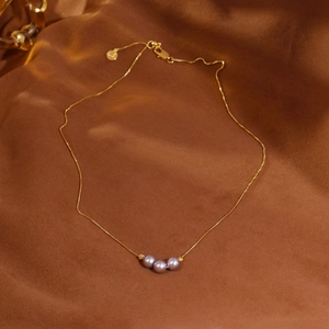 Elegante collar de oro con tres perlas lavanda sobre un lujoso fondo de seda marrón oscuro