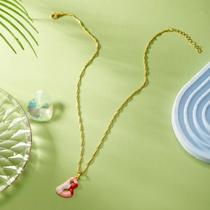 Dettaglio di una collana dorata con pendente a lettera B in smalto rosa su sfondo verde