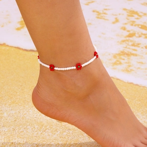 Detail van Boheemse witte kralen enkelband met rode bloemaccenten op een zomerse strandachtergrond