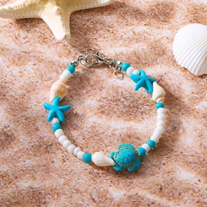 Detail eines Strandarmbands mit blauem Schildkrötenanhänger und Muschelperlen auf Sand
