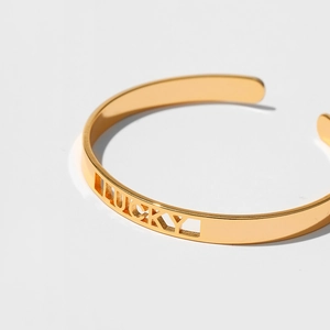 Bracciale rigido dorato minimalista con scritta LUCKY traforata su sfondo grigio pulito